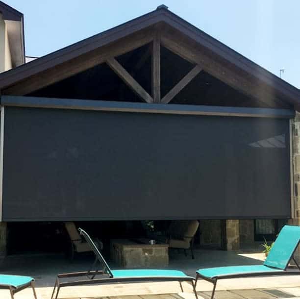 A black outdoor screen is lowered over a patio with turquoise lounge chairs in front, under a wooden roof with stone pillars