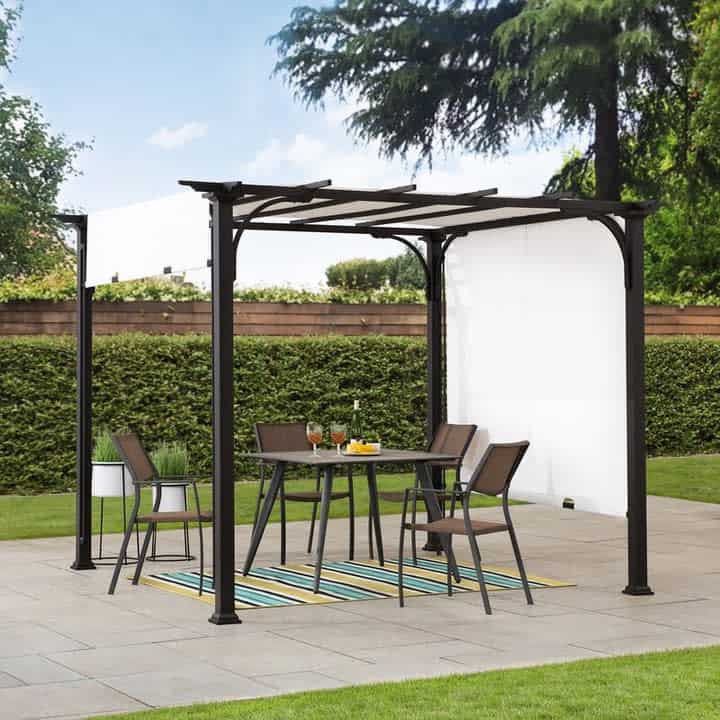 A backyard patio with a retractable pergola, table, and four chairs on a striped rug, drinks are on the table, surrounded by neatly trimmed hedges