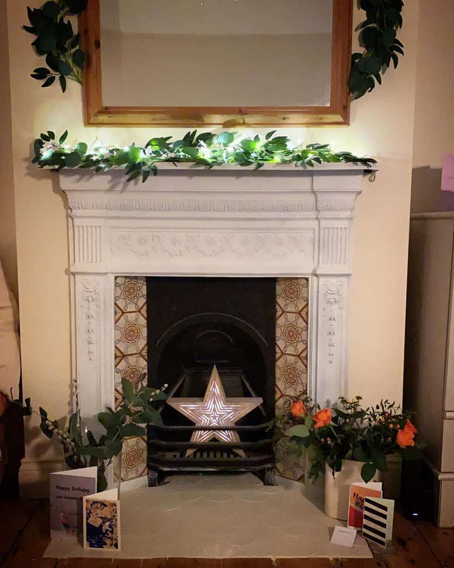 A cozy Victorian fireplace decorated with a garland, star-shaped light, plants, and birthday cards, framed by a wooden mirror for a charming, personal touch