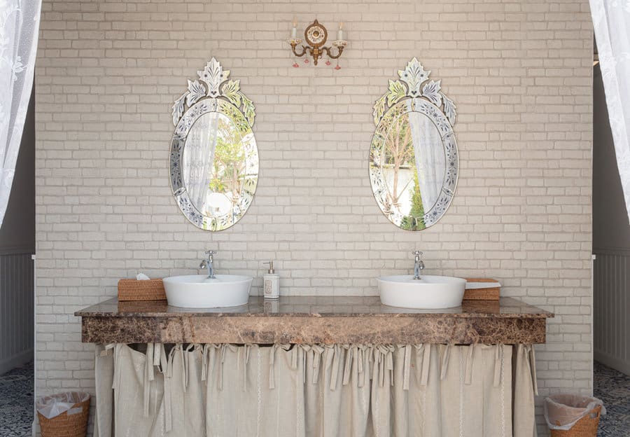 Elegant bathroom with two oval mirrors, white brick wall, double sinks on a marble counter, and soft draped curtains