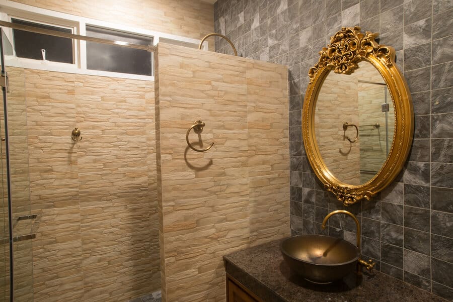 Bathroom with stone walls, a round ornate gold mirror, a bowl sink on a dark counter, and a glass-enclosed shower