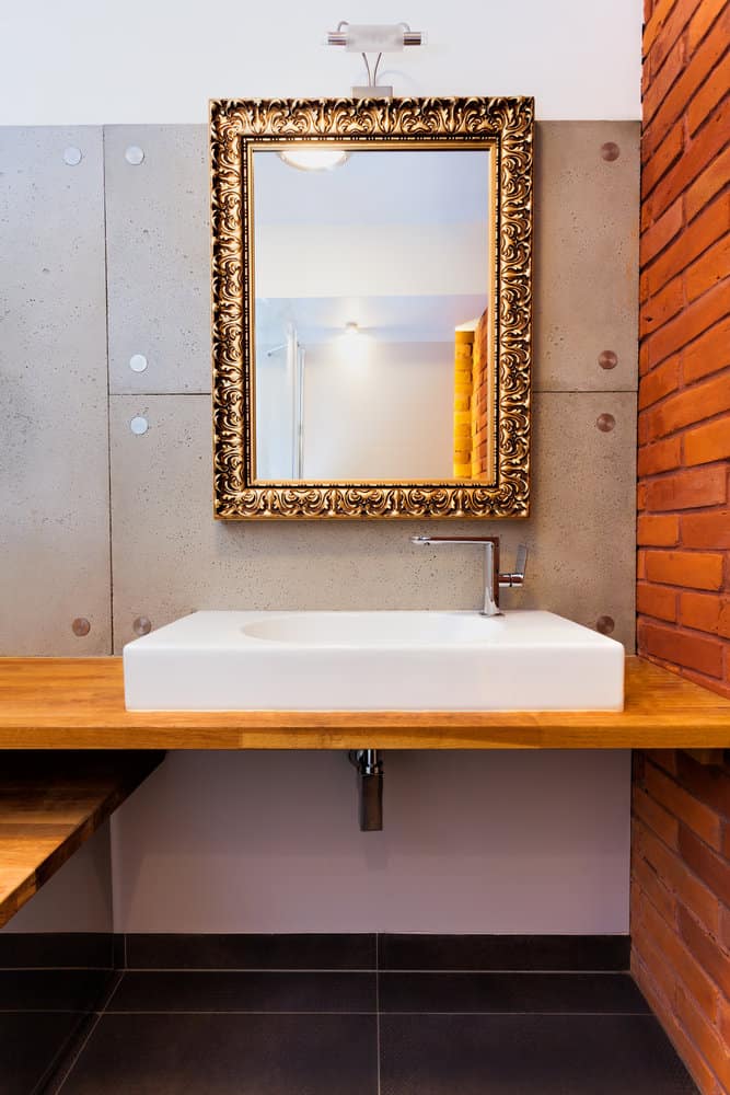 Bathroom with a gold-framed mirror, white rectangular sink, wooden countertop, brick accent wall, and gray tile wall backdrop