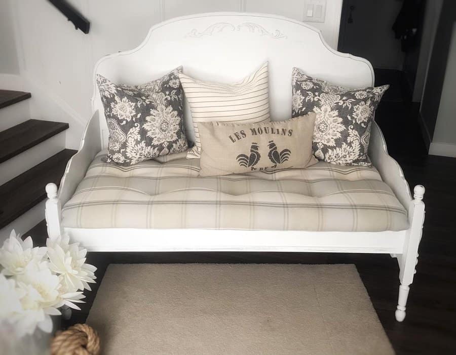 White vintage-style daybed with plaid cushion, floral and rooster-themed pillows, and stairs nearby on dark wood floor