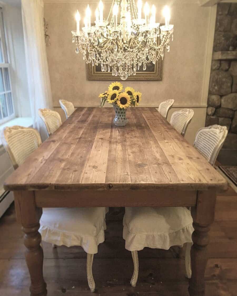 Rustic farmhouse dining room with a wooden table, six white chairs, a vase of sunflowers, and a crystal chandelier above. Cozy and elegant decor