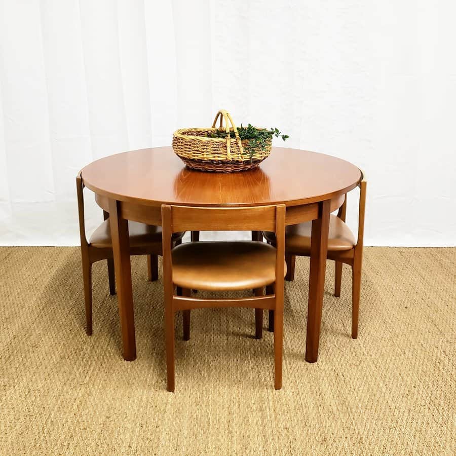 Round wooden dining table with four chairs and a wicker basket with greenery on top, set on a beige carpeted floor