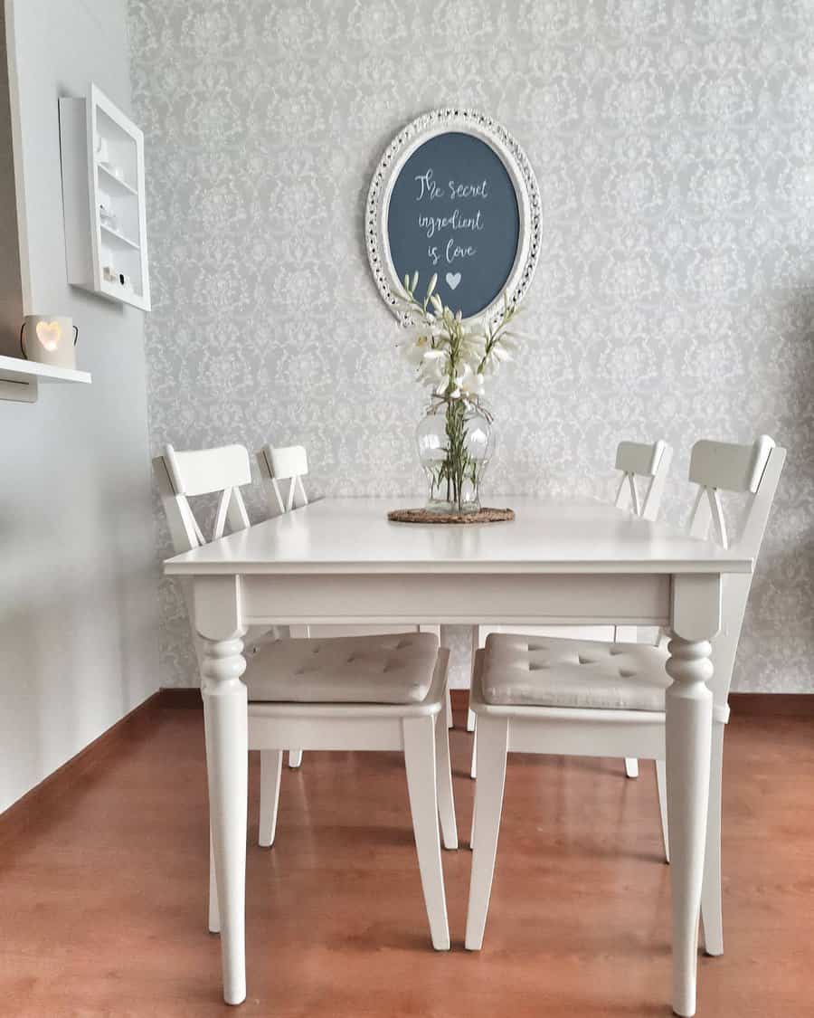 A white dining table with four chairs, a vase with flowers, and a framed chalkboard on a patterned wall reading “The secret ingredient is love”