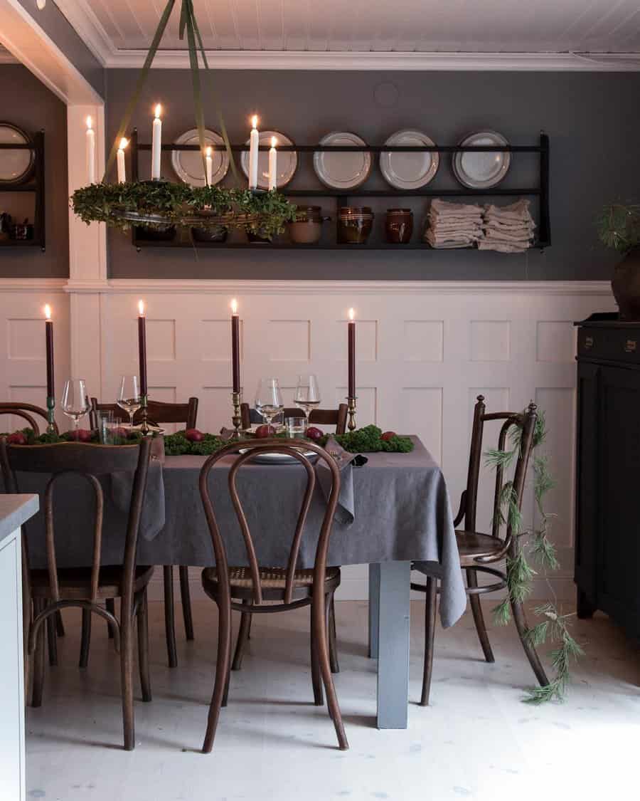 Dining room with a table set for a meal, featuring tall candles, greenery, and wooden chairs. Plates and cups are on wall shelves