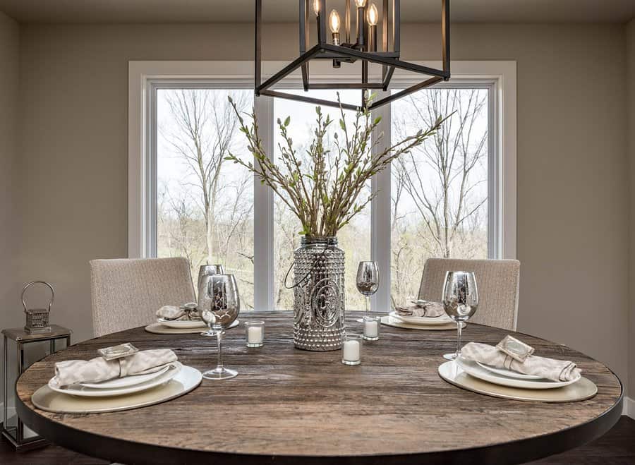 A round wooden dining table set with plates, napkins, wine glasses, and a decorative vase centerpiece near a large window with a view