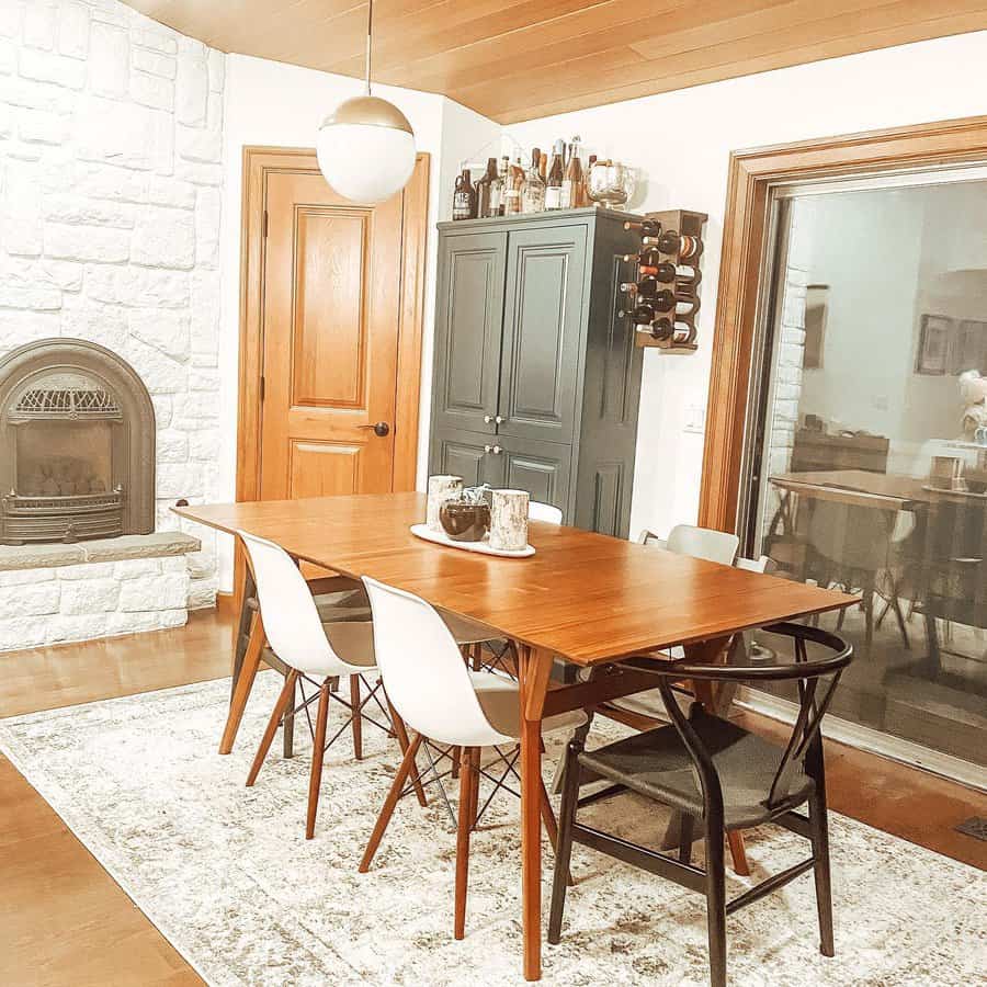 A vintage dining room with a wooden table, white and black chairs, a white stone fireplace, a wine rack, and a cabinet