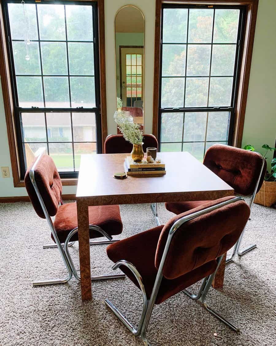 Dining area with a square table, vintage brown chairs, books, and flowers; large windows and a tall mirror in the background