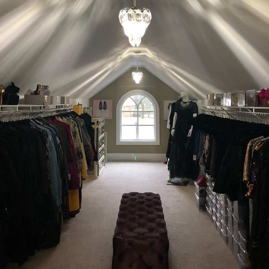 Elegant attic walk-in closet with chandeliers, clothing racks, storage bins, and a tufted bench, creating a boutique-style dressing space.