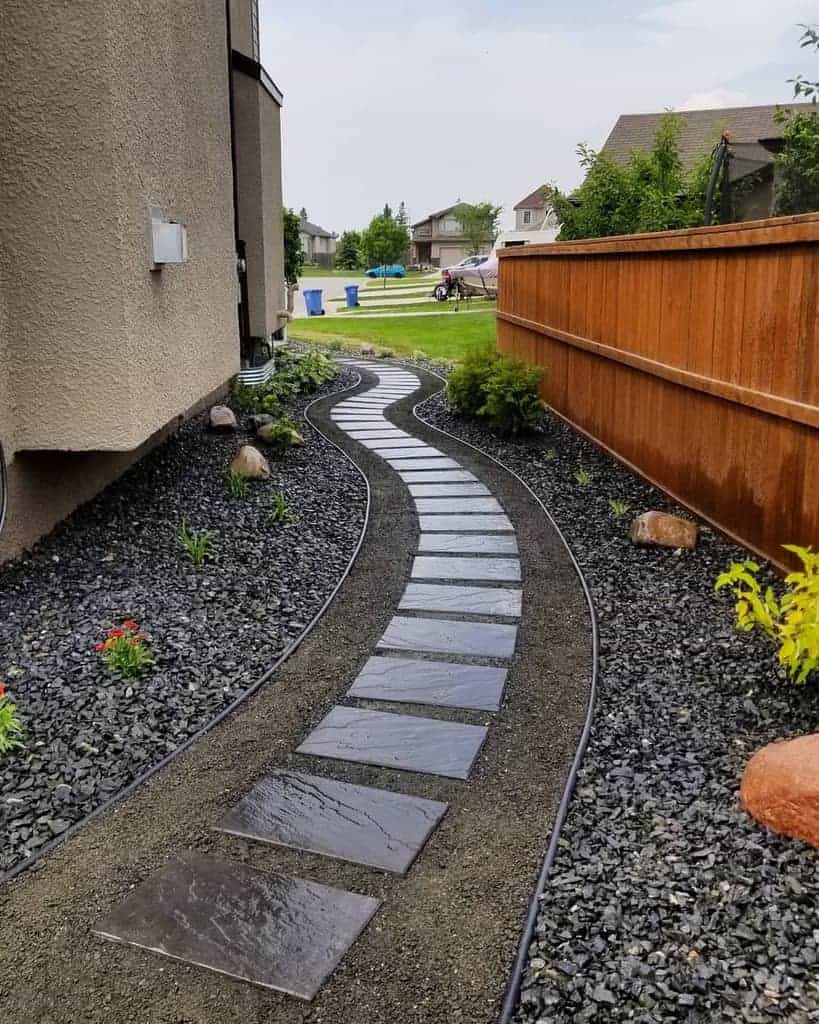 Curved stone path with black gravel on both sides, bordered by a wooden fence and a beige building, leading to a residential area