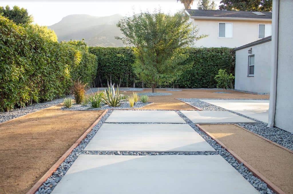 Minimalist garden with square concrete slabs, gravel paths, and a tree in the center, surrounded by green hedges and a white house