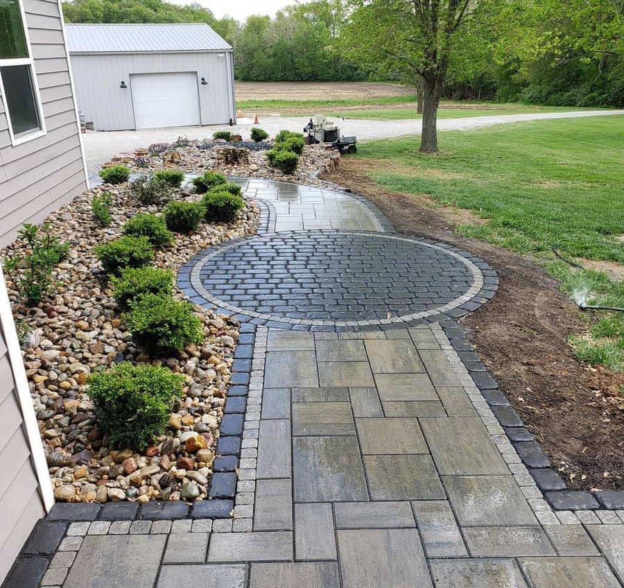 Stone pathway with circular design, surrounded by shrubs and rocks, leading to a garage and green lawn in the background