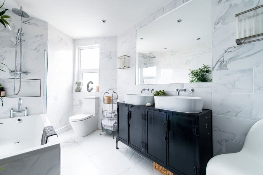 Modern bathroom with marble walls, large mirror, dual sinks on a black vanity, bathtub, and a window; bright and spacious