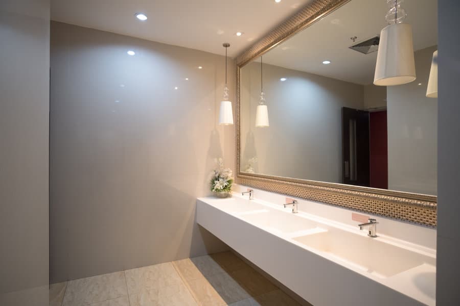 Modern restroom with three white sinks, large mirror, hanging lights, and a small floral arrangement on the counter