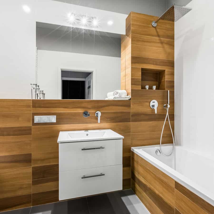 Modern bathroom with wooden paneling, a white sink, bathtub with showerhead, and a large mirror. Soft lighting creates a cozy atmosphere