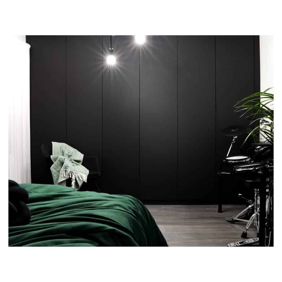 Moody black bedroom with sleek panel walls, emerald green bedding, hanging bulbs, and a corner drum set for a stylish, creative touch