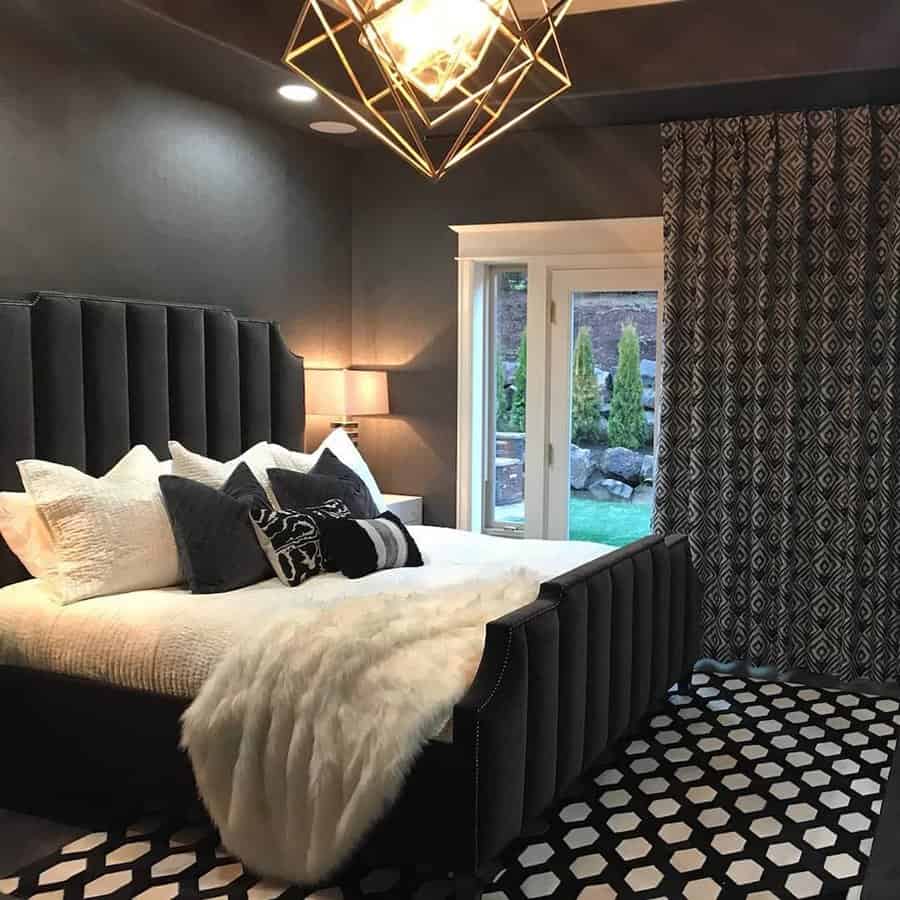 Modern bedroom with dark walls, geometric chandelier, patterned rug, and sliding doors with curtains; black bed with white pillows