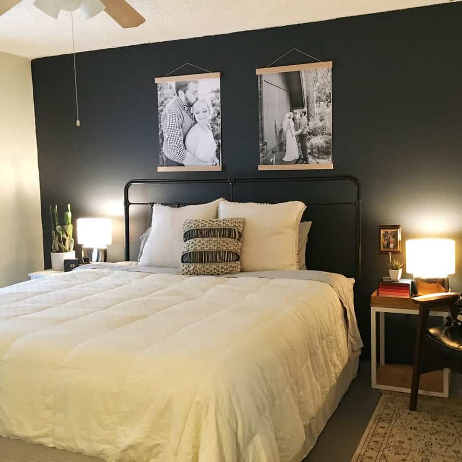 A bedroom with a black accent wall, bed with white bedding, framed photos above, side table with lamps, and a chair to the right