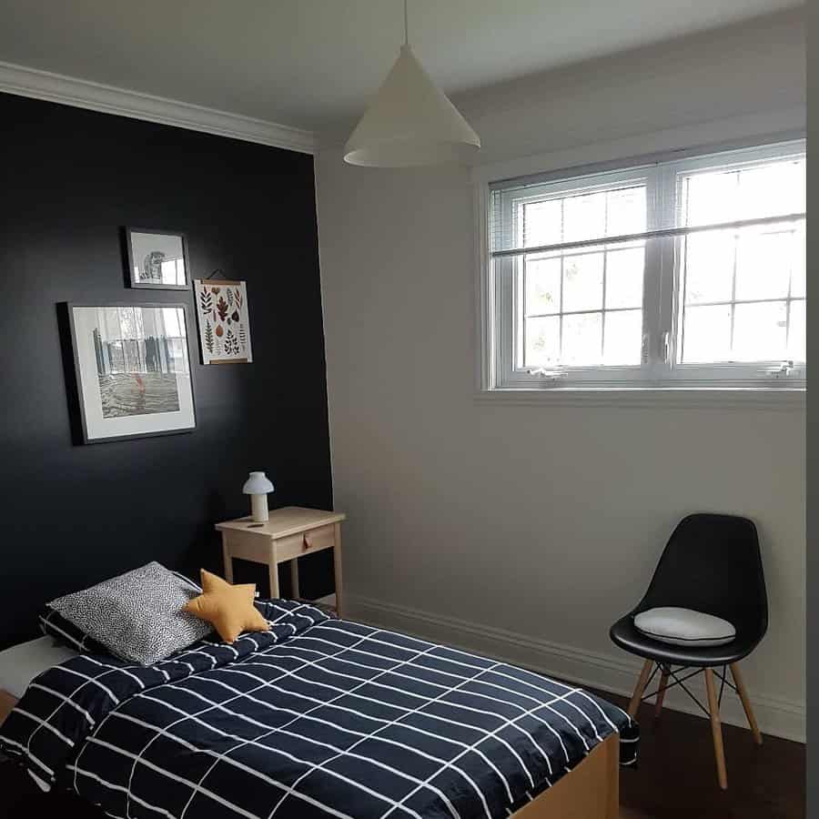 Minimalist bedroom with single bed, black accent wall, framed art, small wooden table with a lamp, and a black chair by the window