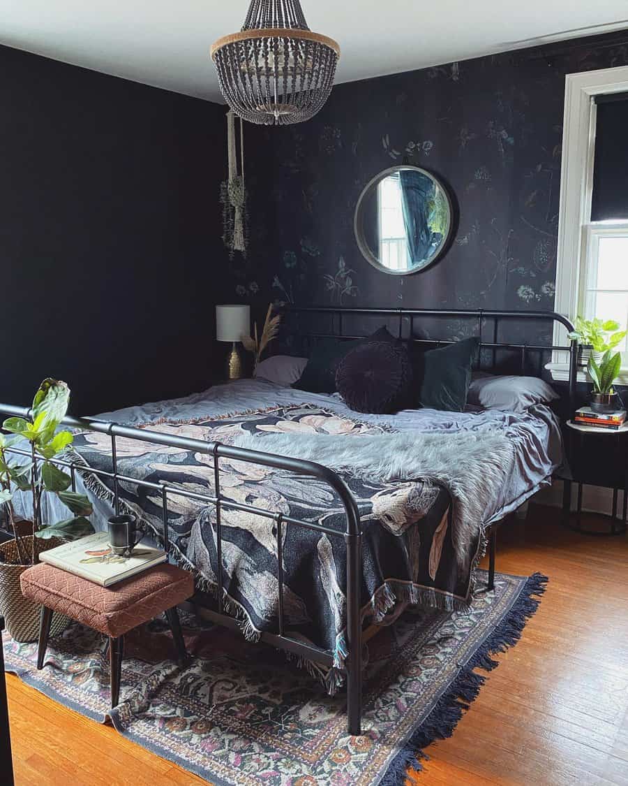 Dark, cozy bedroom with a round mirror, chandelier, patterned rug, and plants; black walls and bedding create a dramatic ambiance