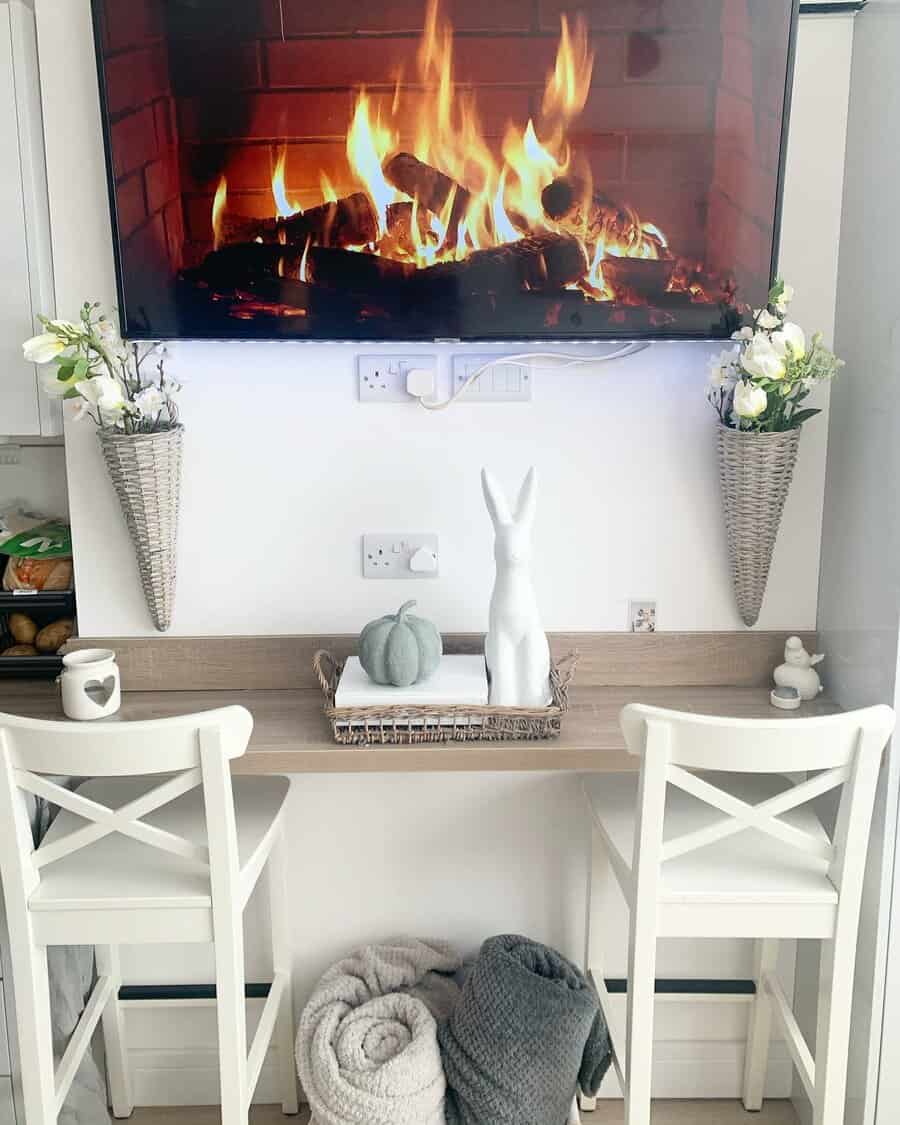 Cozy kitchen nook with two white chairs, a flat-screen TV displaying a fireplace, decorative plants, and a soft blanket on the floor