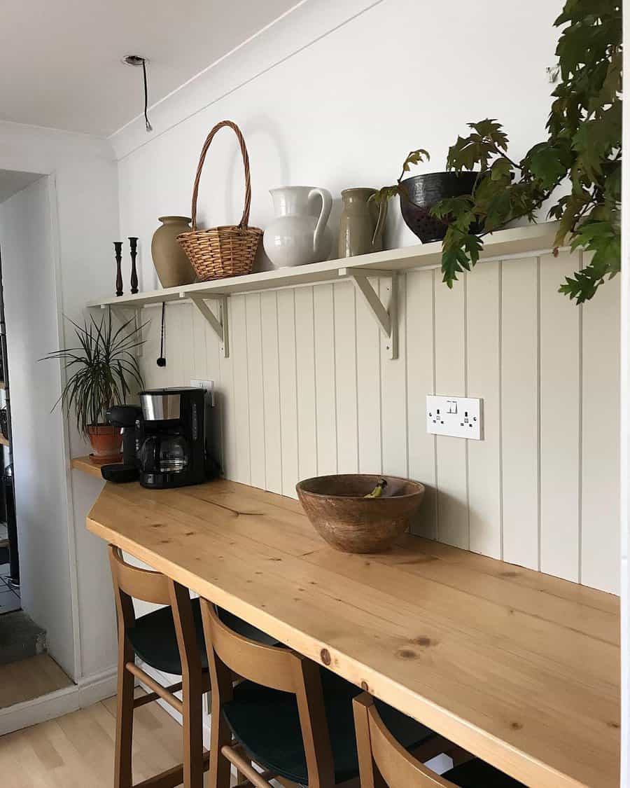 Cozy farmhouse-style breakfast bar with a natural wood countertop, simple wooden chairs, beadboard wall paneling, and rustic decor on open shelving