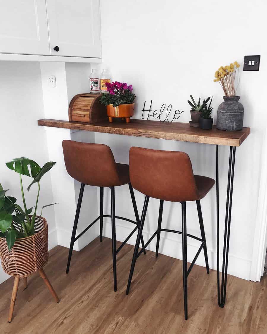 Minimalist kitchen nook with two brown chairs at a wooden bar table, plants, a bread box, and decor items on top against a white wall