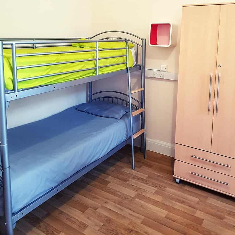 A room with a metal bunk bed, green bedding on top, blue on bottom, a wooden wardrobe, and a small wall shelf