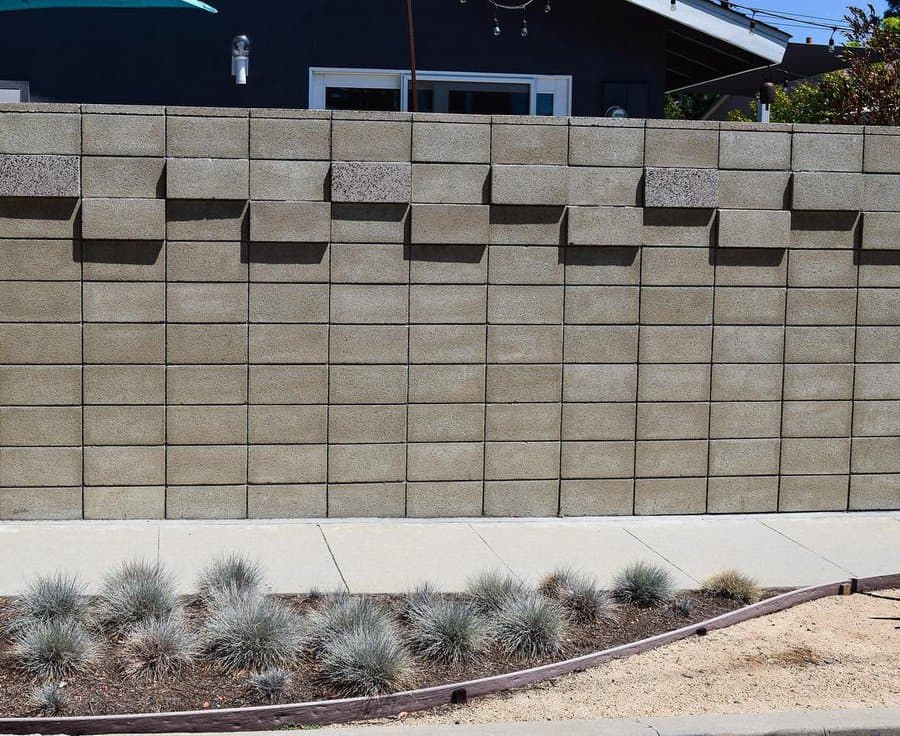 A concrete block wall with a geometric pattern, in front of a house, bordered by a sidewalk and ornamental grasses