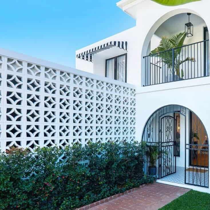 Modern white house with arched doorway, patterned cinder block wall, black balcony, and green hedge under a clear blue sky