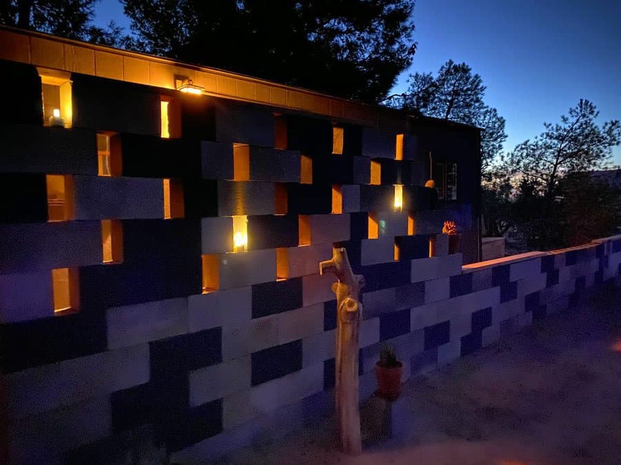 A cinder block wall with glowing lights at dusk, surrounded by trees against a fading blue sky, creates a serene atmosphere