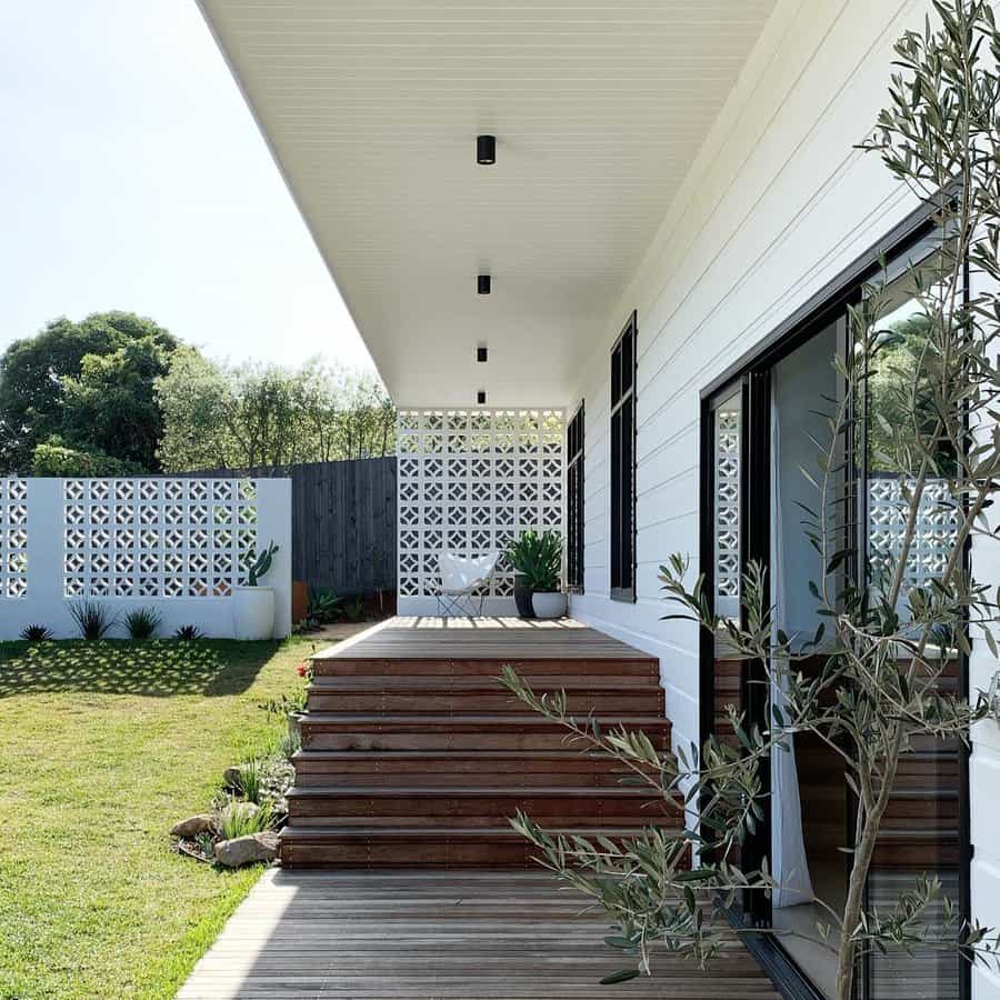 Modern house with a wooden deck, white exterior cinder block walls, and decorative block fence, steps lead to a grassy yard with plants