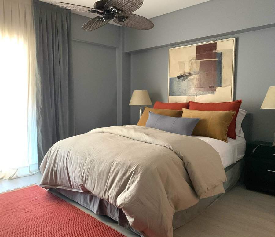 Cozy bedroom with a beige bed, colorful pillows, abstract artwork, and a ceiling fan; light curtains and floor lamp create a serene vibe