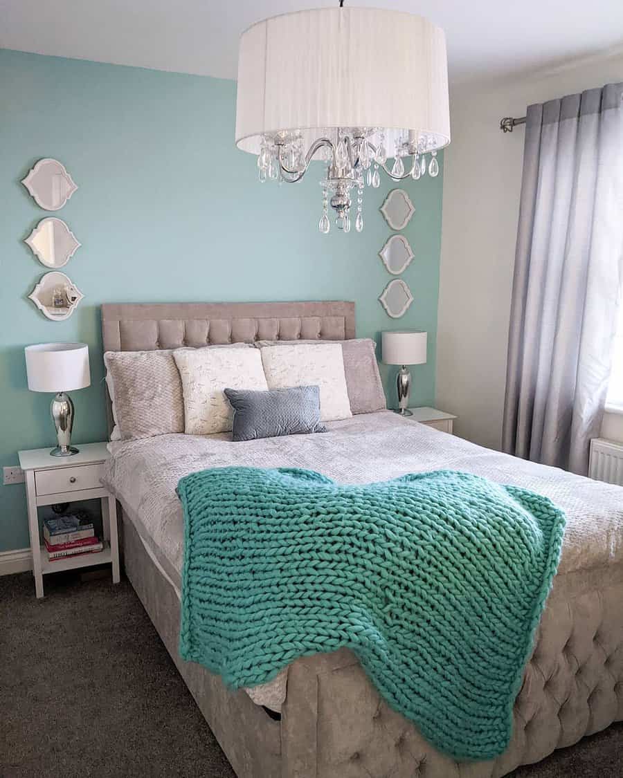 Cozy bedroom with a tufted bed, mint green accent wall, chandelier, white lamps, mirrors, and a chunky teal throw blanket