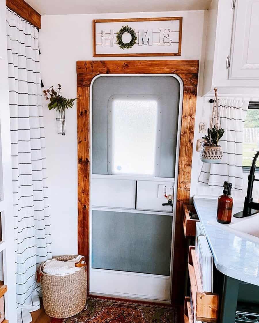 A cozy RV entryway with a rustic "HOME" sign, plants, a woven basket, and a door framed in natural wood, adding a welcoming and homely feel to the space.