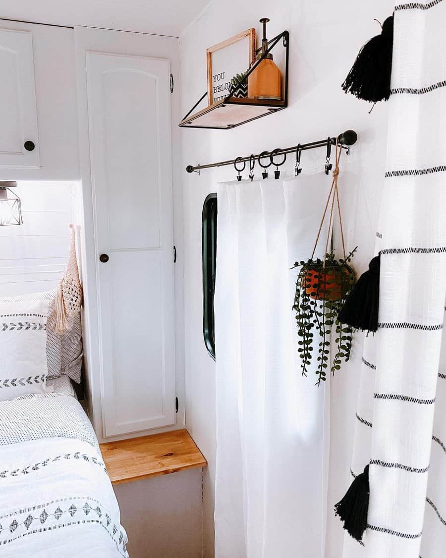 A charming RV bedroom corner featuring minimalist decor, a small shelf with a framed sign, hanging plants, and a cozy bed, creating a welcoming and stylish atmosphere.