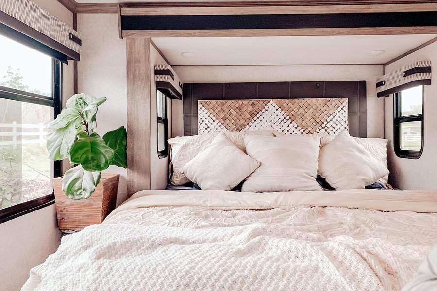 A serene RV bedroom with a cozy bed, textured headboard, neutral pillows, and a large plant, creating a peaceful and inviting space with natural light.