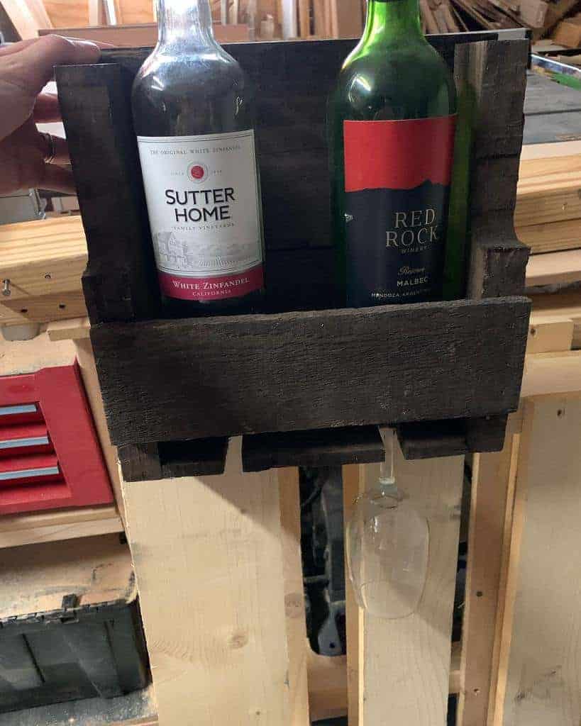 Wooden shelf holding two wine bottles and an upside-down hanging wine glass, set against a cluttered workshop background