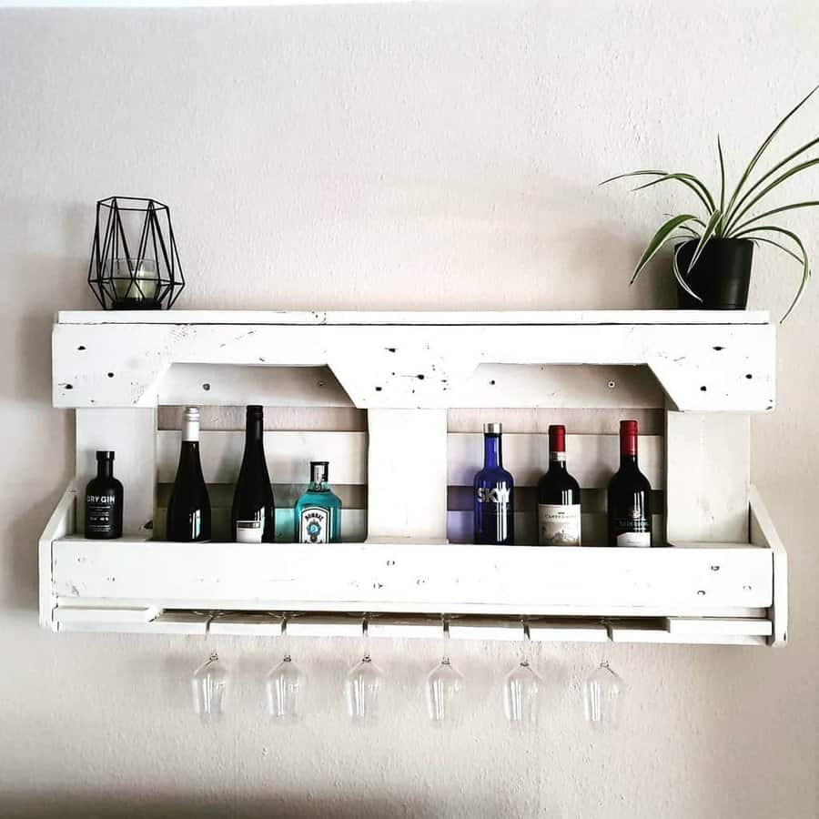 White wooden wall shelf holding liquor bottles and glassware, with a geometric black decor piece and potted plant on top