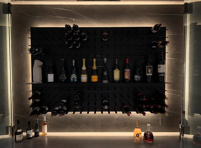 Wall-mounted wine rack with various bottles, underlit for highlighting; additional bottles are placed on a counter beneath