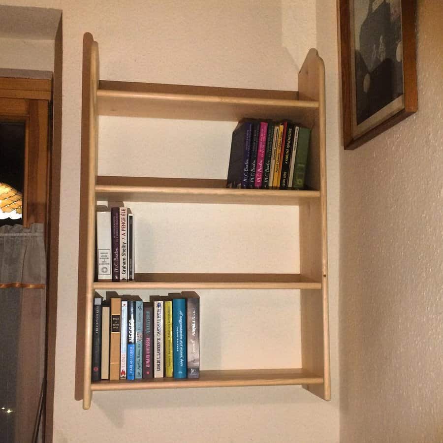 Simple wooden wall-mounted bookshelf with a minimalist design, holding a small collection of books in a cozy room with warm lighting.