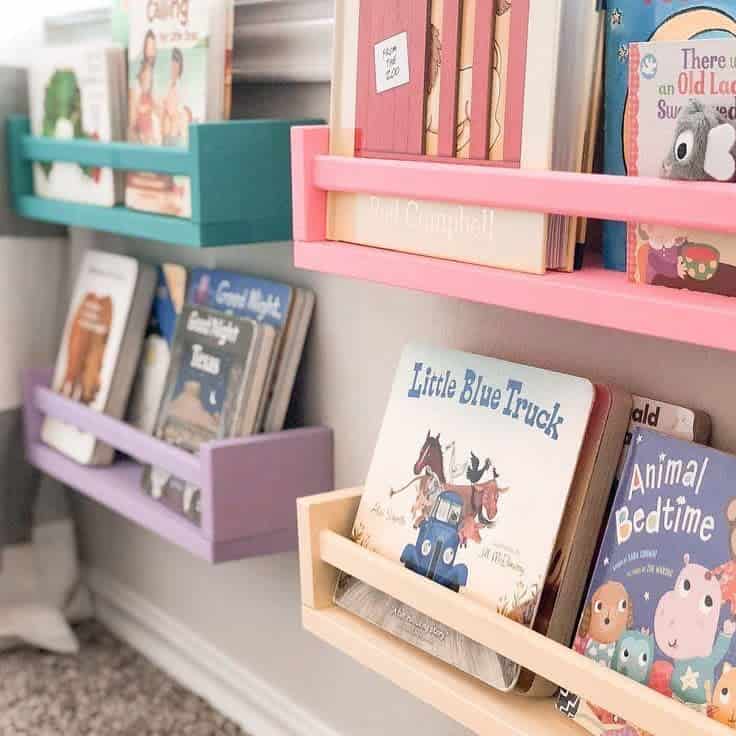 Colorful wall-mounted bookshelves at a child's level, neatly holding board books in pastel pink, purple, teal, and beige racks.