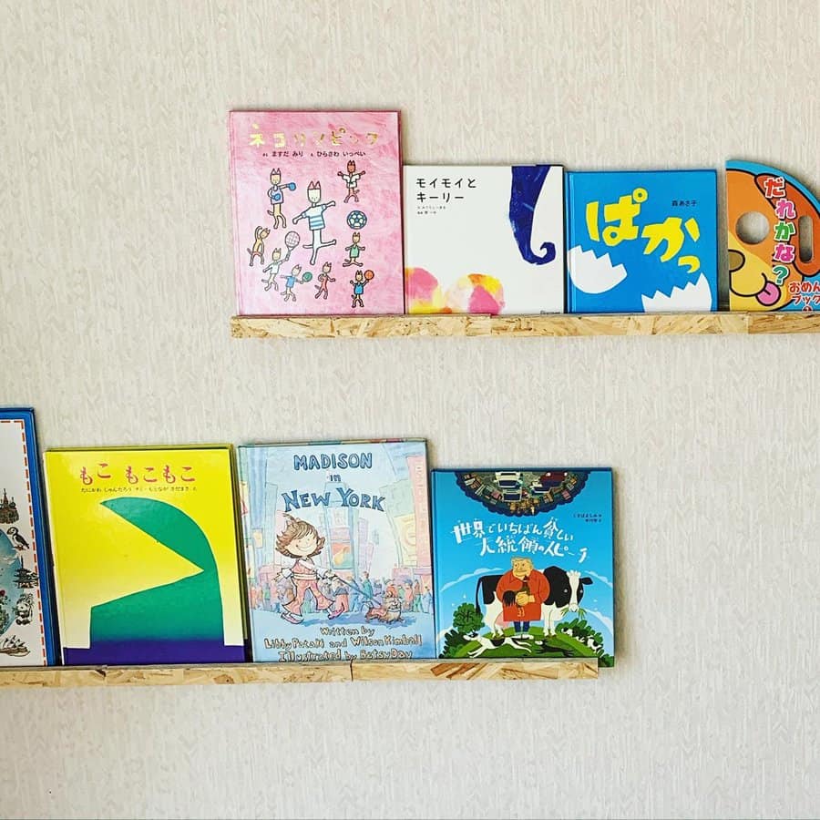 Minimalist wall-mounted wooden shelves displaying colorful children's books in various languages, arranged neatly on a light-textured wall.