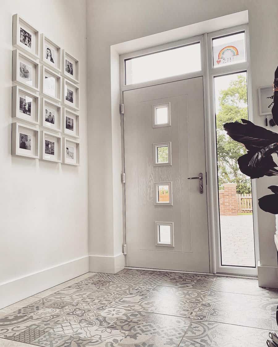 A bright entryway with a white door featuring small windows, patterned tile flooring, and a wall displaying a grid of framed photos