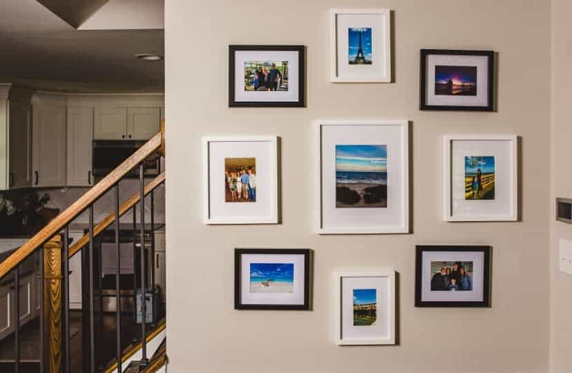 A wall display of various framed photos, arranged in a grid, beside wooden staircase railing in a home interior