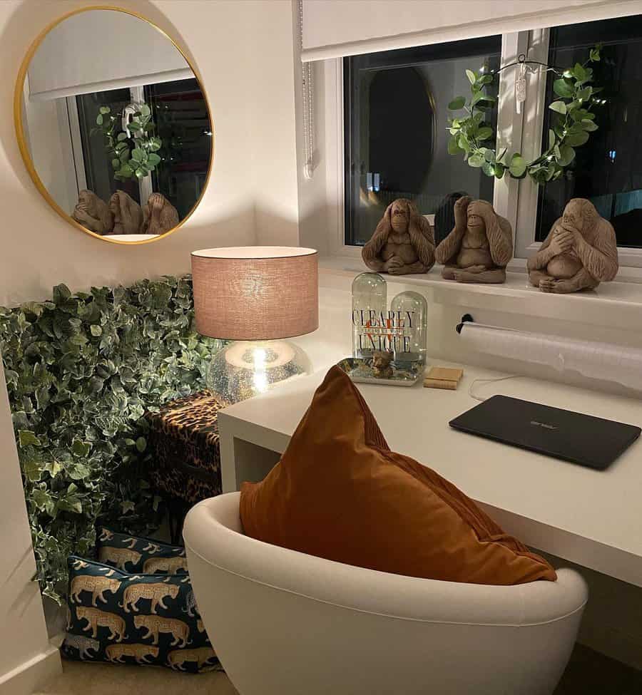 Cozy desk with a laptop, lamp, and decorations, including a "speak no evil" monkey trio, next to a round mirror and leafy plants