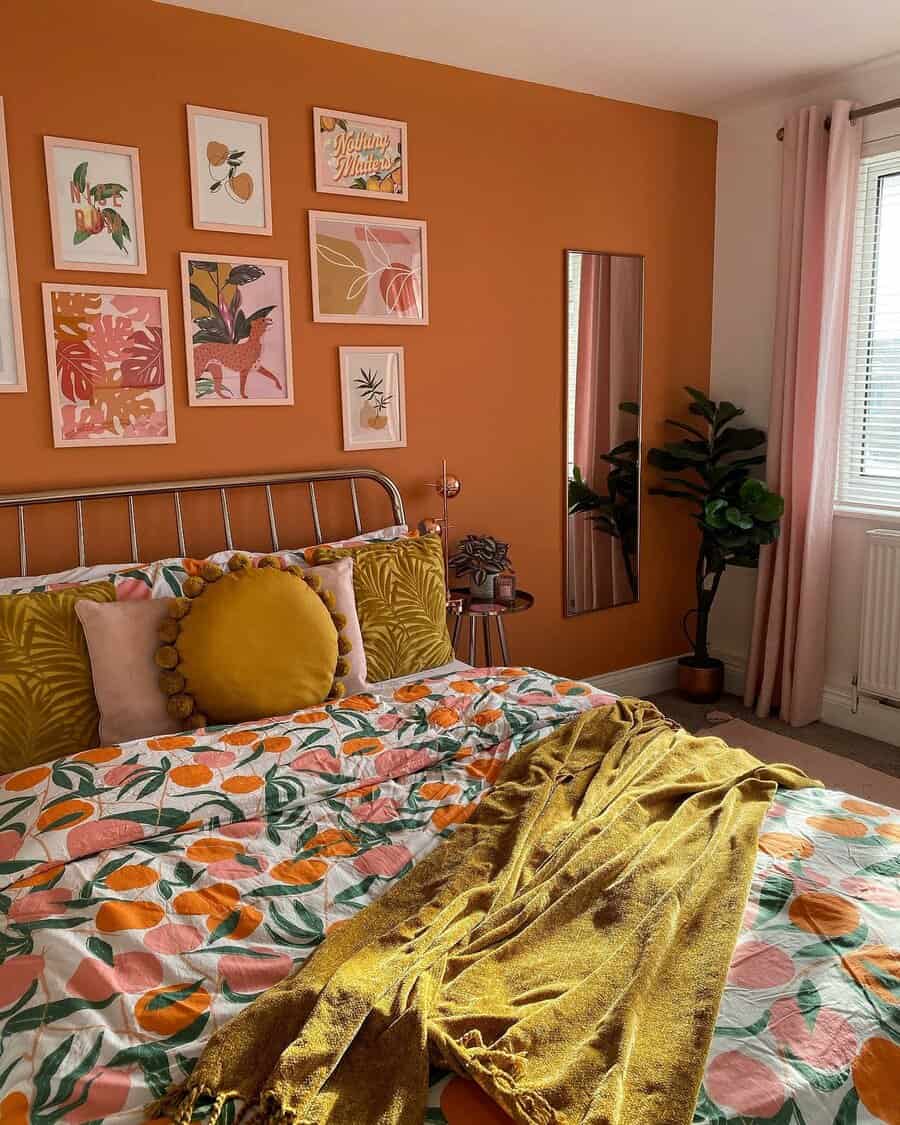 Warm bedroom with orange accent wall, vibrant floral bedding, gallery wall art, and a large potted plant; mirror beside the bed