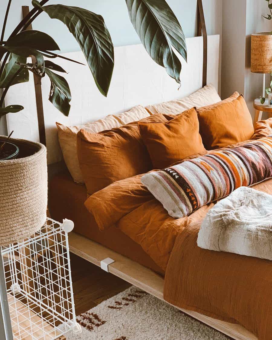 Cozy warm bedroom with orange bedding, a striped pillow, and large green plants. A textured lamp is on a small wooden table beside the bed.